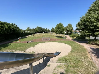 Wassererlebnisspielplatz Bruchhausen-Vilsen Blick über einen großen Wasserspielplatz mit Grünflächen, Bäumen und einer langen Edelstahlrutsche.