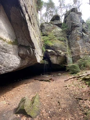 Diebshöhle Hohnstein Felsformation mit kleinem Wasserfall, umgeben von Bäumen und Moos, auf einem mit Laub bedeckten Waldboden.