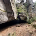 Diebshöhle Hohnstein Felsformation mit kleinem Wasserfall, umgeben von Bäumen und Moos, auf einem mit Laub bedeckten Waldboden.
