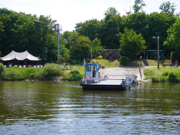 Bar Hol-üBar Eine kleine Fähre überquert einen ruhigen Fluss, umgeben von üppigem Grün und einem Veranstaltungszelt.