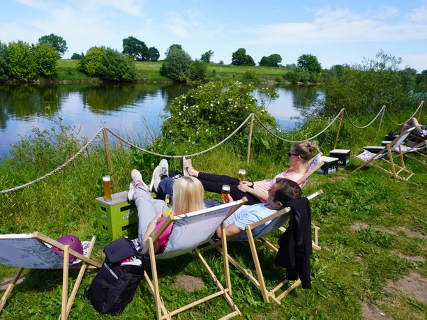 Bar Hol-üBar Drei Personen entspannen an einem Flussufer auf Sonnenliegen, umgeben von grüner Natur und blauem Himmel.