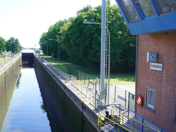 The picture shows the Drakenburg lock with water channel, surrounded by green areas and a brick building.