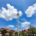 Maibaum auf dem Marktplatz Ein Maibaum steht auf einem Marktplatz, flankiert von Gebäuden und umrahmt von blühenden Bäumen.