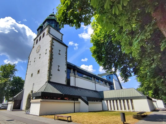 Weiße Kirche mit grünem Turm, flankiert von Bäumen, unter wolkenlosem, blauem Himmel in Hövelhof.