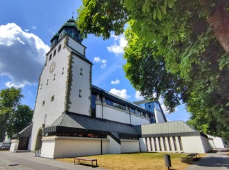 St. Johannes Nepomuk Kirche Weiße Kirche mit grünem Turm, flankiert von Bäumen, unter wolkenlosem, blauem Himmel in Hövelhof.