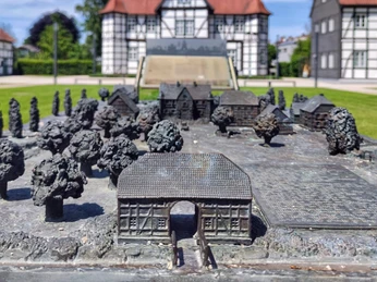 Bronzeplastik eines Schlossgartens vor einem Fachwerkhaus unter blauem Himmel mit weißen Wolken.