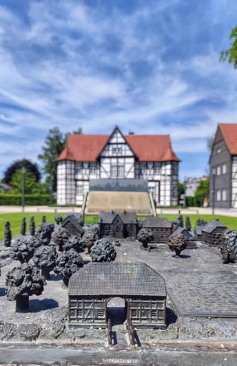 Geländemodell Schlossgarten Bronzeplastik eines Schlossgartens vor einem Fachwerkhaus unter blauem Himmel mit weißen Wolken.