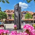 Glockenspiel aus dunklem Stein auf einem Marktplatz, umgeben von Blumen, Bäumen und Fachwerkhäusern.