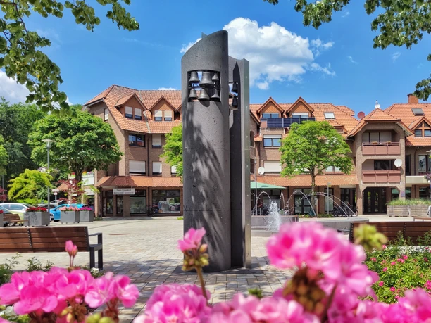 Glockenspiel aus dunklem Stein auf einem Marktplatz, umgeben von Blumen, Bäumen und Fachwerkhäusern.
