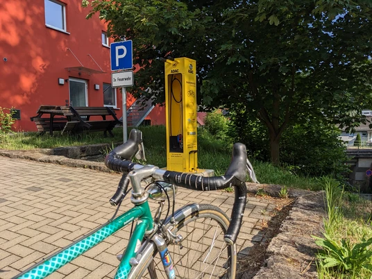 ADAC Fahrradstation Stadt Wehlen Ein grünes Fahrrad steht vor einer gelben Fahrradreparaturstation neben einem roten Gebäude mit einem blauen Parkplatzschild und einem Baum im Hintergrund.