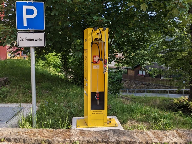 Geel ADAC fietsservicestation naast een blauw parkeerbord met het opschrift "3x Feuerwehr", omgeven door groene bomen en gras.