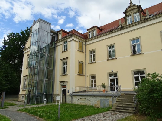 Gedenkstätte Pirna Sonnenstein Gelbes Gebäude mit rotem Dach und modernem Glasaufzug, umgeben von Grünflächen und Bäumen, bei bewölktem Himmel.