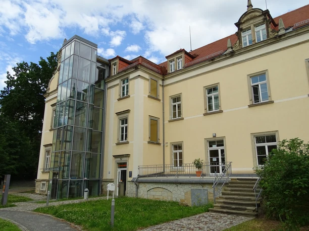 Gedenkstätte Pirna Sonnenstein Gelbes Gebäude mit rotem Dach und modernem Glasaufzug, umgeben von Grünflächen und Bäumen, bei bewölktem Himmel.