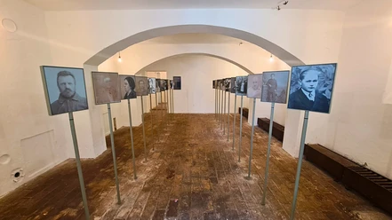 A room with white walls and a vaulted ceiling shows black and white portraits of people mounted on stands and arranged in two rows.