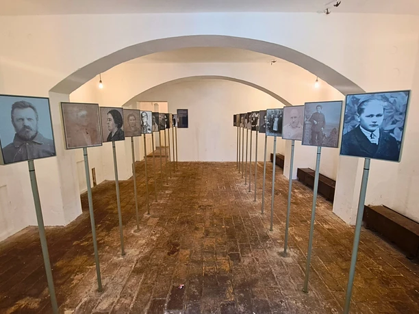 A room with white walls and a vaulted ceiling shows black and white portraits of people mounted on stands and arranged in two rows.