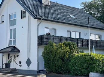 Zweistöckiges Wohngebäude mit dunklem Dach und Balkon, umgeben von grünem Blattwerk.