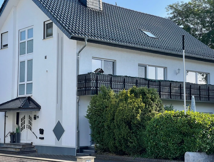 Zweistöckiges Wohngebäude mit dunklem Dach und Balkon, umgeben von grünem Blattwerk.