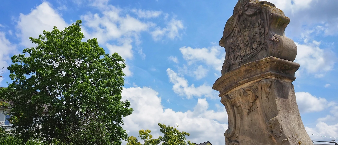 Steinerner Apostelstein mit kunstvollen Gravuren auf einer Grünfläche unter blauem Himmel.