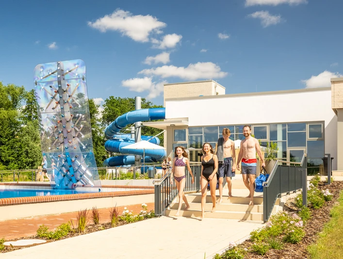 Vier Menschen gehen zu einem modernen Freibad mit Rutsche und Kletterwand bei sonnigem Wetter.