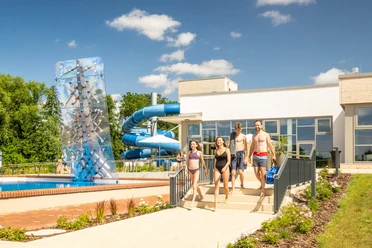 Freizeitbad Aqua Fun Kirchlengern Vier Menschen gehen zu einem modernen Freibad mit Rutsche und Kletterwand bei sonnigem Wetter.