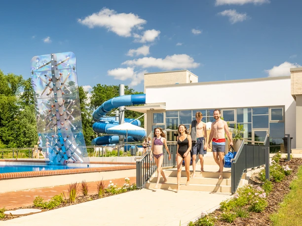 Freizeitbad Aqua Fun Kirchlengern Vier Menschen gehen zu einem modernen Freibad mit Rutsche und Kletterwand bei sonnigem Wetter.