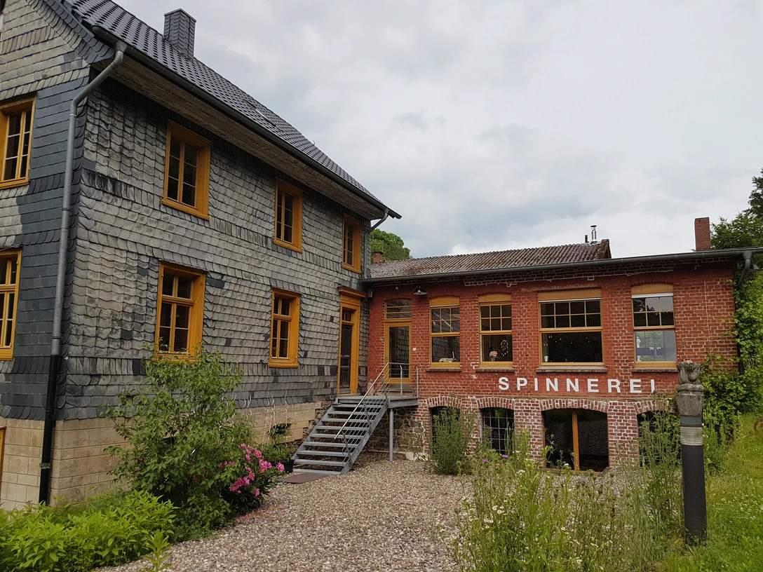 Spinnerei Historisches Gebäude mit Schieferfassade, modernem Anbau und „Spinnerei“-Schriftzug im Grünen.