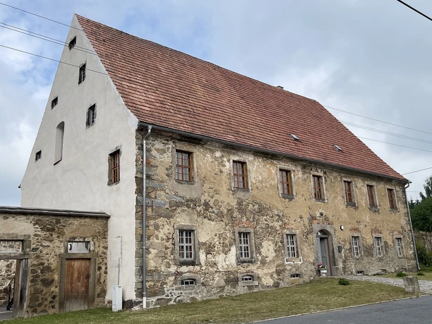 Old stone house with a red tiled roof and several small windows, surrounded by a green meadow and a gray sky in the background.