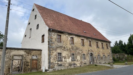Gesindehaus Polenz Altes Steinhaus mit rotem Ziegeldach und mehreren kleinen Fenstern, umgeben von grüner Wiese und grauem Himmel im Hintergrund.