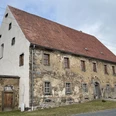 Gesindehaus Polenz Altes Steinhaus mit rotem Ziegeldach und mehreren kleinen Fenstern, umgeben von grüner Wiese und grauem Himmel im Hintergrund.
