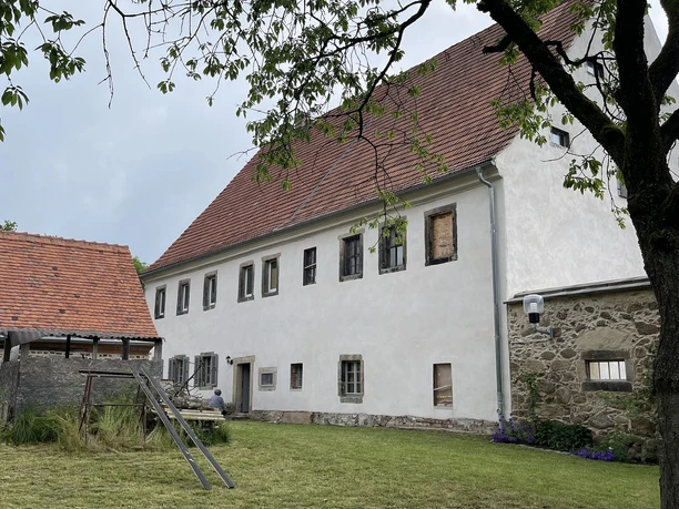 Gesindehaus Polenz Rückseite mit Milchhaus Weißes Gebäude mit rotem Ziegeldach, umgeben von grünem Rasen und einem Baum im Vordergrund; ruhige ländliche Atmosphäre.
