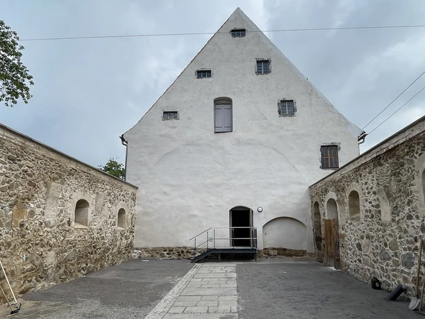 Gesindehaus Polenz, Innenhof mit Pferdestall Historisches Gebäude mit weißer Fassade und steinernen Seitenmauern, umgeben von einem gepflasterten Hof; bewölkter Himmel im Hintergrund.