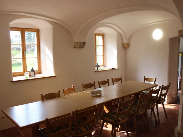 Gesindehaus Polenz Kaminzimmer Heller Raum mit einem langen Holztisch und zehn Stühlen, zwei Fenster lassen Tageslicht herein, auf dem Tisch steht ein Schild mit der Aufschrift "Stammtisch".