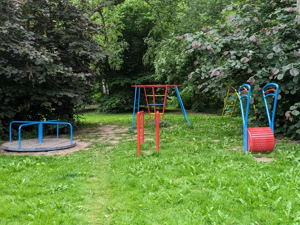 Natucamp an der Weser Spielplatz Waldspielplatz mit Wippe, Klettergerüst und Drehscheibe, umgeben von grünen Sträuchern und Bäumen.