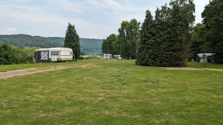 Wohnwagen stehen auf einer grünen Wiese neben Bäumen, mit bewaldeten Hügeln im Hintergrund.