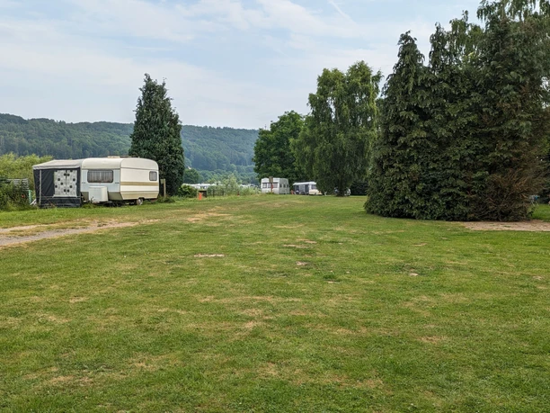 Natucamp an der Weser Wiese Wohnwagen stehen auf einer grünen Wiese neben Bäumen, mit bewaldeten Hügeln im Hintergrund.