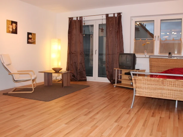 IMG_1145.JPG Ein helles Wohnzimmer mit Holzboden, gemütlicher Sitzecke, dezenter Beleuchtung und großen Fenstern.