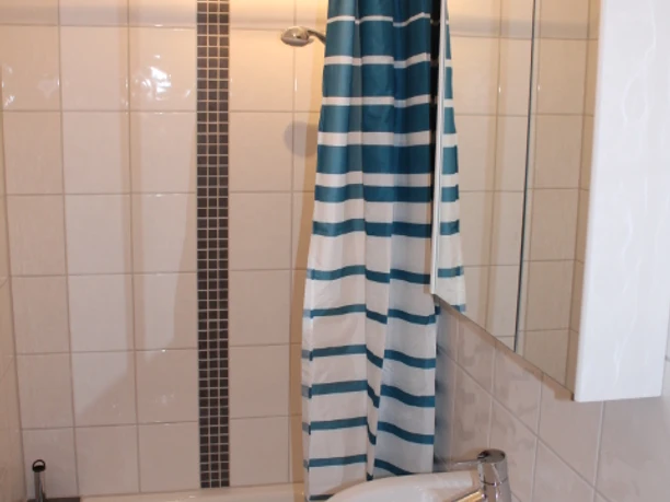 IMG_1150.JPG Duschkabine mit gemusterter Duschvorhang neben einem weißen Waschbecken, gefliest in Weiß mit grauer Streifenakzent.