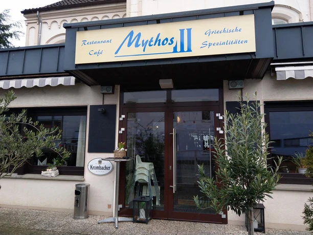 Eingang des griechischen Restaurants Mythos mit großem Schild und Pflanzen vor der Tür