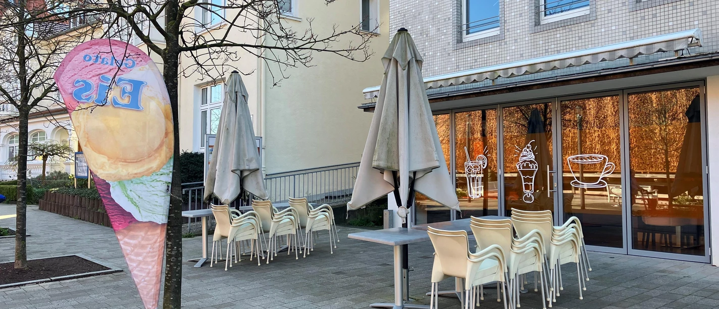 Eiscafé Therme: Außenbereich mit gestapelten Stühlen, Sonnenschirmen und Eisfahne