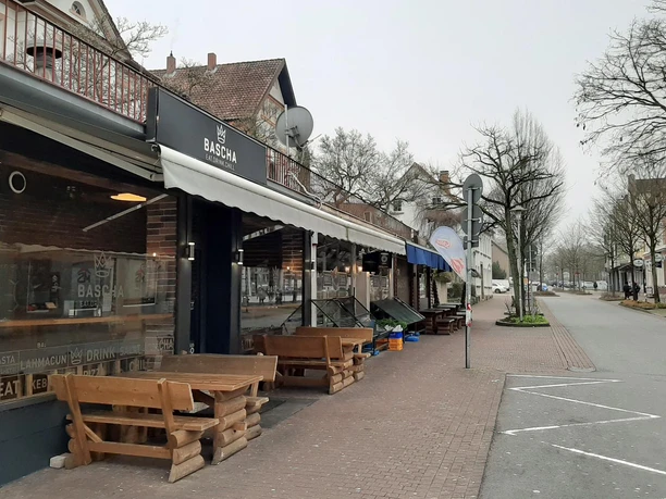 Außenansicht des Bascha mit schwarzer Markise, Holzbänken und ruhiger Straße