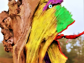 Herr von Schwindebeck Ein bemalter Baumstumpf zeigt ein farbig gestaltetes Gesicht mit bunten gelben, grünen, violetten und roten Zügen, die einem Kunstwerk ähneln, inmitten einer Landschaft.