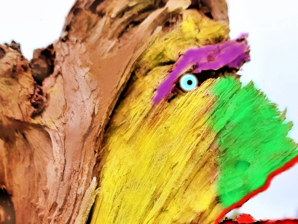 Ein bemalter Baumstumpf zeigt ein farbig gestaltetes Gesicht mit bunten gelben, grünen, violetten und roten Zügen, die einem Kunstwerk ähneln, inmitten einer Landschaft.