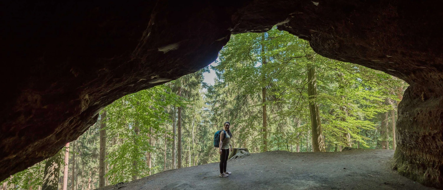 Diebskeller Diebshöhle Eine Person mit Rucksack steht am Eingang einer dunklen Höhle, umgeben von einem grünen Wald bei Tageslicht.