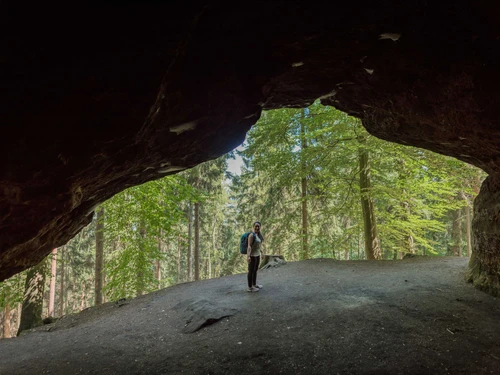 Eine Person mit Rucksack steht am Eingang einer dunklen Höhle, umgeben von einem grünen Wald bei Tageslicht.