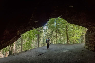 Thieves' cellar Thieves' den A person with a rucksack stands at the entrance to a dark cave, surrounded by a green forest in daylight.