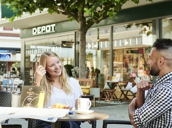 Zwei Personen sitzen an einem Tisch im Freien vor einem Café, umgeben von Geschäften und Bäumen.