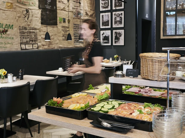 Ein reichhaltiges Frühstücksbuffet mit Auswahl an Aufschnitt, Gemüse und Obst in einem modernen Café.