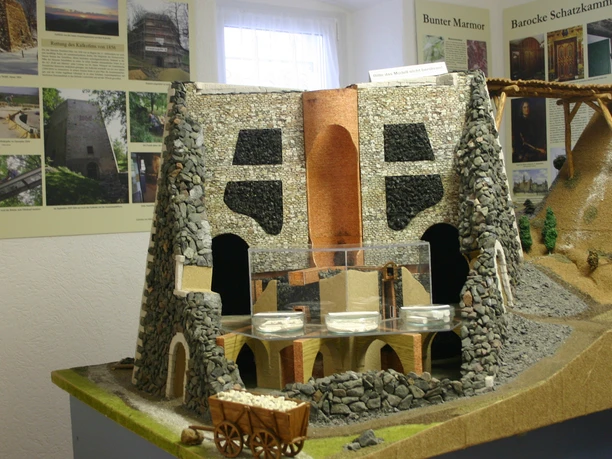 Model van een historische kalkoven met stenen muren en een kleine houten wagen vol stenen, omringd door informatieborden op de muur.