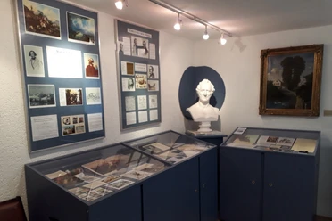 A small exhibition room with blue showcases, a white bust, framed pictures and documents on the walls, brightly lit.