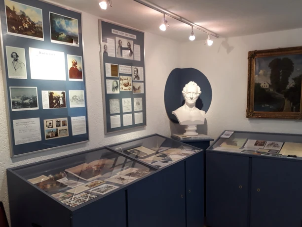 Een kleine tentoonstellingsruimte met blauwe vitrines, een witte buste, ingelijste foto's en documenten aan de muren, helder verlicht.